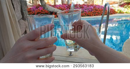 Two Hands Holding Glasses With White Vine Together Over Blue Pool In Luxury Hotel Room Surrounded By