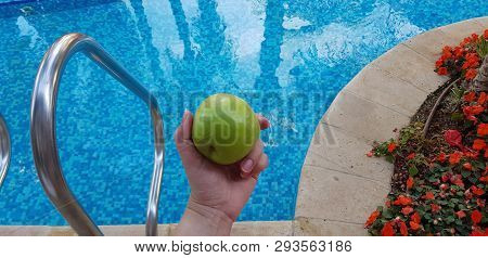 Green Apple In Female Hand Over Blue Waters Of Outdoor Swimming Pool In The Summer
