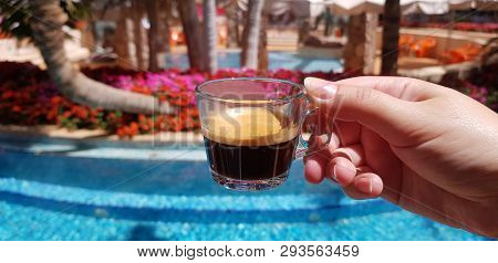 Small Coffee Cup Made From Transparent Glass In Female Hand Against Blue Outdoor Pool Surrounded By 