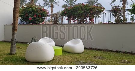Three White Plastic Chairs Stay Empty Round A Small Table On The Grass Under Palm Tree In Hotel Gard