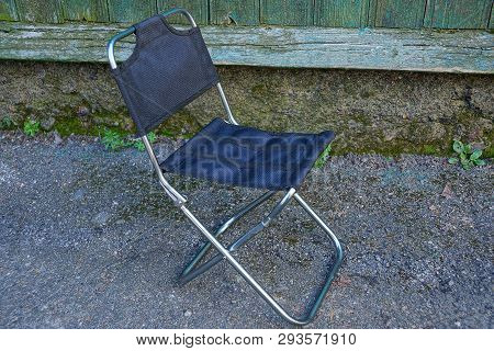 A Small Folding Metal Chair With Black Matter Stands On The Asphalt Against A Wall Outside