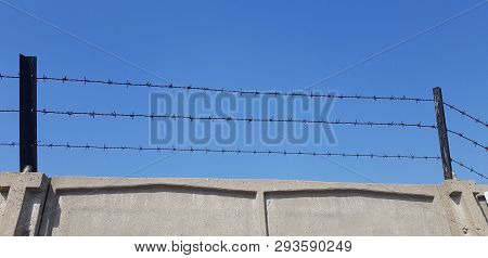 Three Rows Of Barbed Wire Over Gray Concrete Fence Against Blue Skies At Background