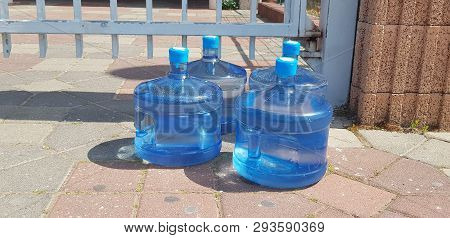 Four Big Plastic Jars Full With Drinking Water Stand On The Ground Near Metal Fence