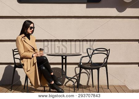Take Away Coffee In Women's Hands. Beautiful Young Brunette Woman Wearing Brown Coat Sitting At Tabl