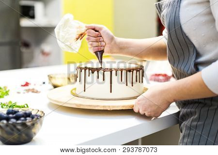 A Confectioner Squeezes Liquid Chocolate From A Pastry Bag Onto A White Cream Biscuit Cake On A Wood