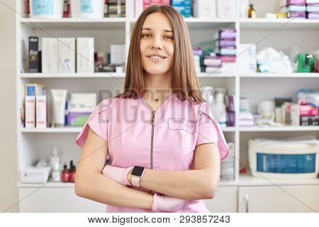 Positive Nice Young Beautician Stands In Her Cabinet With Folded Hands And Pose And Looks At Camera.