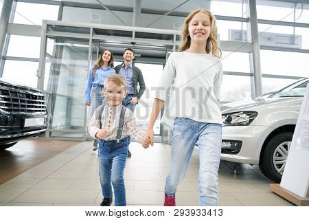 Front View Of Funny Little Boy With Elder Sister Entering In Big Auto Showroom. Cute Children With P