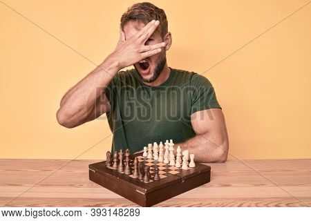 Young caucasian man playing chess sitting on the table peeking in shock covering face and eyes with hand, looking through fingers afraid 