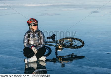 Woman Sitting Near Her Bike On Ice. The Girl Cyclist Stopped To Rest. She Sits On The Wheel And Enjo