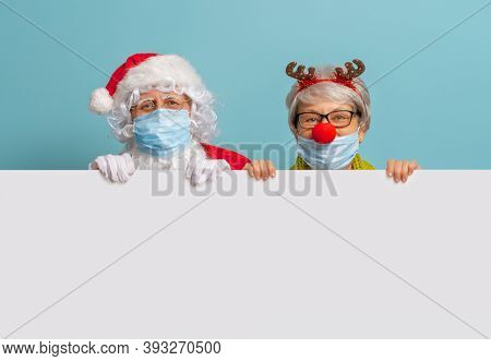 Merry Christmas and Happy Holidays! Santa Claus and reindeer on bright color background. Senior people in carnival costumes wearing face masks during Covid-2019
