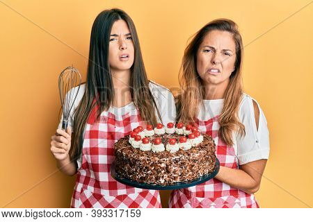 Hispanic family of mother and daughter wearing baker apron holding homemade cake clueless and confused expression. doubt concept. 