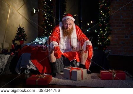 Bad Evil Santa Claus With A Red Beard Sits On The Bed And Collects Gifts On Christmas Eve. A Man In 