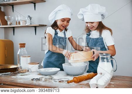 Two Little Girls In Blue Chef Uniform Working With Flour By Using Sieve On The Kitchen.