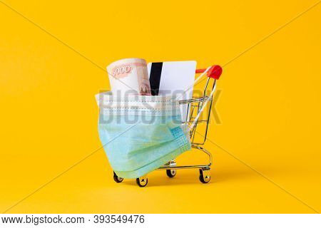 A Shopping Cart With A Medical Mask On, Filled With Cash And A Bank Card. Yellow Background. Close U