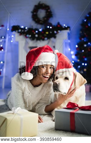 African American Girl In Santa Hat Stroking Labrador Dog While Lying On Floor Near Gift Boxes On Blu