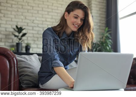 Smiling Young Woman Sitting On Sofa With Laptop Computer And Chating With Friends.