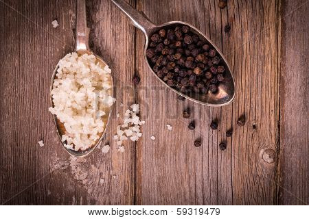 Vintage effect image of tarnished silver spoons filled with salt crystals and black peppercorns, over rough wood background. Space for your text