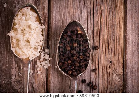 Vintage effect image of tarnished silver spoons filled with salt crystals and black peppercorns, over rough wood background. Space for your text