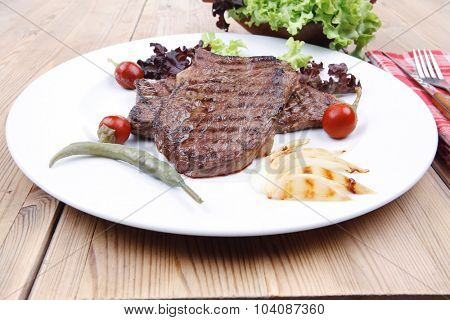 meat food : two grilled steak on green lettuce salad , with roast onion and red hot chili peppers , on dish over wooden table