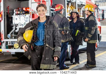 Portrait of happy firewoman with male colleagues discussing by truck in background at fire station