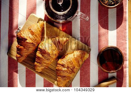 Three French Freshly Baked Croissants On Woode With Teapot Honey Herbs And Knife On Striped Cloth