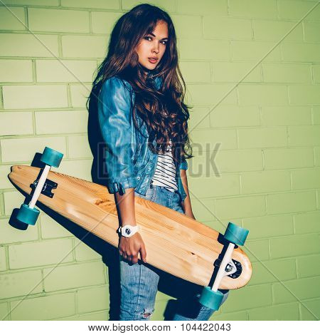 Beautiful Long-haired Girl With A Wooden Long Skateboard Near A Green Brick Wall