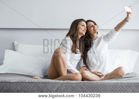 Two Hot Girls Lying  On A Bed Taking A Photo Of Themselves With A Phone