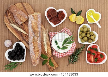 Picnic food with french baguette loaf on an olive wood board, camembert cheese, olives, sun dried and fresh tomatoes, oil, lemon, garlic and herb leaf sprigs.