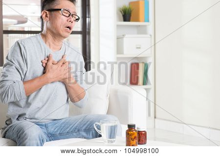 Mature 50s Asian man heartache, pressing on chest with painful expression, sitting on sofa at home, medicines on table.
