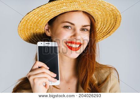 A young red-haired woman in a hat and with red lipstick shows a black screen of the phone in the camera, a mosk up and smiles