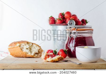 A kitchen table scene of freshly made strawberry jam in storage jar with a tin pail of strawberries in the background; bread butter and strawberries on a wooden chopping board.