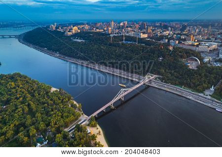 Panoramic view of a modern city with a river. Skyline bird eye aerial view of the old part of the city under dramatic cloud sunset sky. General view of the right bank of Kiev with the Dnieper River, a pedestrian bridge and a large park area at sunset. Kie
