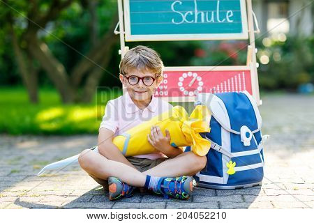 Happy little kid boy with glasses sitting by desk and backpack or satchel. Schoolkid with traditional German school bag called Schultuete on his first day to school. Child outdoors on warm sunny day
