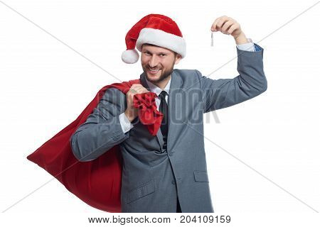 Portrait of happy smiling man in suite and red cap, like santa holding bag with presents over shoulder and showing key with arm up. Young santa clause giving in present house or car for xmas.