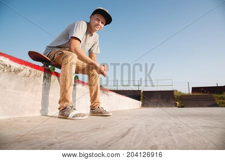 A teenager in a cap sits on a skateboard in the park and smiles