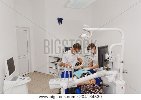 The male dentist with a female assistant helps to treat the teeth of a woman patient in a clinic in the office