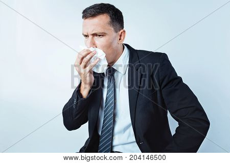 Got sick. Waist up shot of a frustrated man of business looking into vacancy while using a napkin and sneezing over the light background.