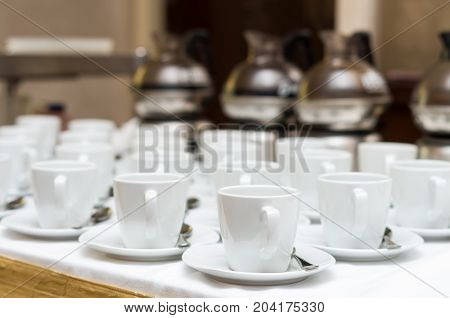 Many rows of white coffee cup with saucer and teaspoon on table and coffee maker background