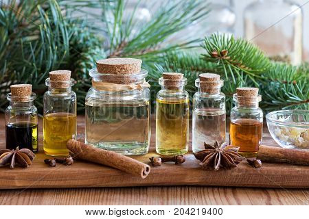 Selection of essential oils with spruce and pine branches on a wooden background with Christmas spices and ingredients in the foreground: cinnamon clove star anise frankincense