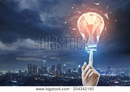 Female hand pointing at abstract polygonal lamp on night city background with cloudy sky and copy space. Innovation concept