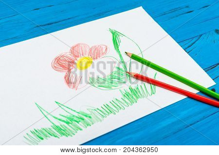 The red and green crayons lie on the drawn flower picture on blue wooden background