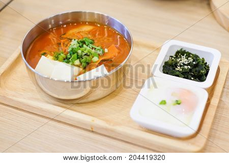 The homemade food set consists of spicy kimchi soup (bacon slice pork white tofu spring onion & cabbage) soft boiled egg (onsen) green seaweed with white sesame serve on wooden tray.
