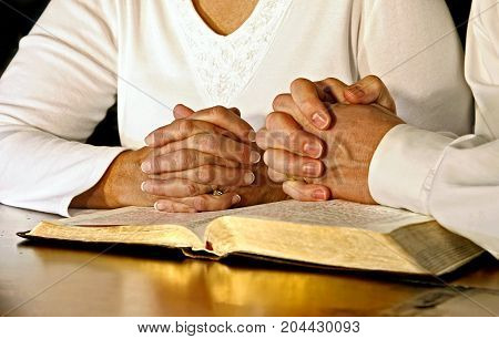 A married couple wearing white shirts clasp their hands in prayer together over an open Holy Bible. Main focus point is on the woman's hands.