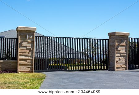 Black metal driveway entrance gates set in sandstone brick fence with residential garden in background