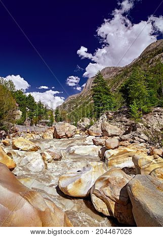The Ganges River flows across the town. Houses in rocky river valley and green forest against snow capped mountain in the distant. Panoramic view of river. Gangotri. Uttarakhand. India.