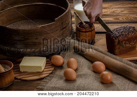 The hand cuts black bread on the background of ingredients for cooking bread. eggs honey milk butter flour sieve rocking chair for dough on a background of burlap and wooden background