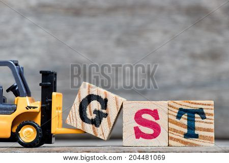 Yellow plastic forklift hold letter G to complete word GST (Abbreviation of Goods and Services Tax) on wood background