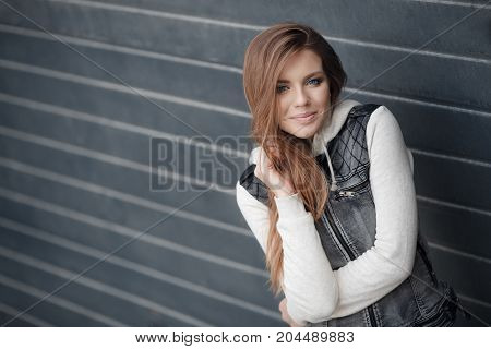 Cute girl with blue eyes,long, straight,light brown hair,beautiful smile,dressed in a knitted pullover blue-gray and gray jacket with no sleeves,minimal makeup,spends time alone in the city,posing against the backdrop of the gray wall,spring portrait