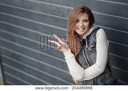 Cute girl with blue eyes,long, straight,light brown hair,beautiful smile,dressed in a knitted pullover blue-gray and gray jacket with no sleeves,minimal makeup,spends time alone in the city,posing against the backdrop of the gray wall,spring portrait