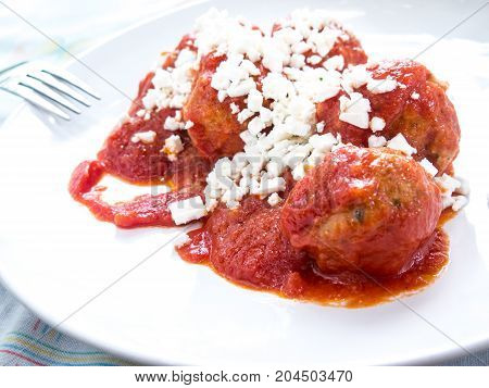 Some meatballs on a white plate in a red tomato sauce and some hard ricotta cheese on top